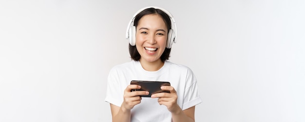 Happy asian girl gamer playing on mobile phone watching on smartphone wearing headphones standing over white background