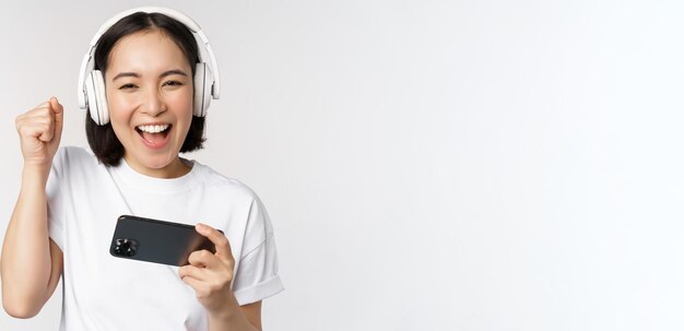 白い背景の上に立っているヘッドフォンを身に着けているスマートフォンで見ている携帯電話で遊んで幸せなアジアの女の子ゲーマー