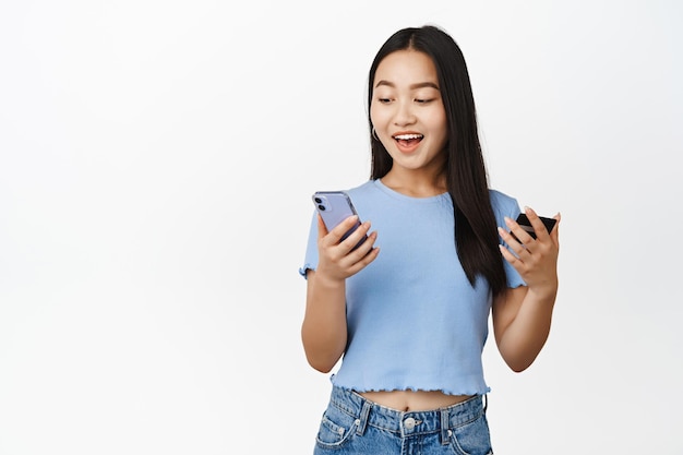 Free photo happy asian girl holding credit card and mobile phone looking at smartphone screen excited purchase online in app white background