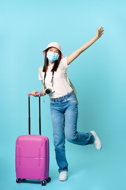 休暇旅行に行くスーツケースに興奮してポーズをとる医療マスク観光客の幸せなアジアの女の子 無料の写真