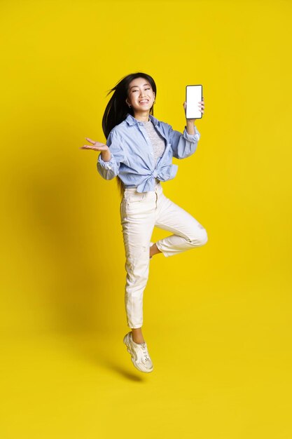 Happy asian girl jumping holding smartphone showing white screen mobile app advertisement isolated on yellow background Product placement