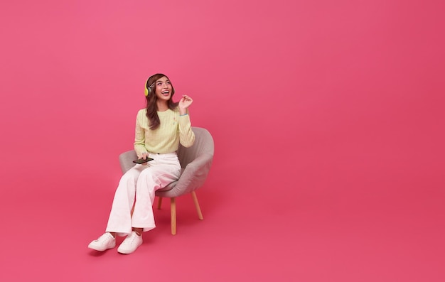 Happy Asian girl listening favourite music playlist on moblie phone application with wireless headphones on chair isolated on pink copy space studio background