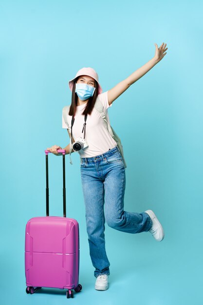 의료용 마스크를 쓴 행복한 아시아 소녀가 여행가방을 들고 흥분한 포즈를 취하고 있다.