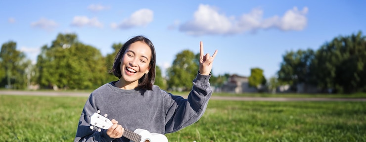 無料Photo 楽しいアジアの女の子が公園でウクレレを弾いて ロック・アンド・ロール・ヘビー・メタル・ホーンを示し 笑顔でサインしています