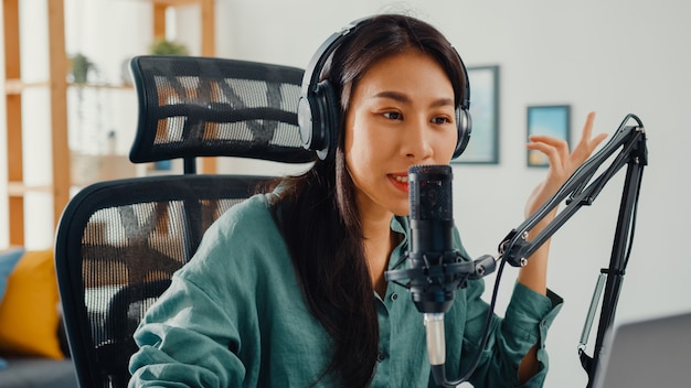 Happy asian girl recording a podcast on her laptop computer with headphones and microphone