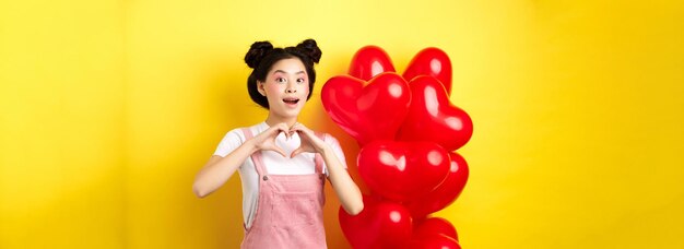 Happy asian girl showing heart sign near romantic balloons waiting for true love on valentines day l