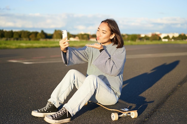 Happy asian girl sits on skateboard takes selfie with longboard makes cute faces sunny day outdoors