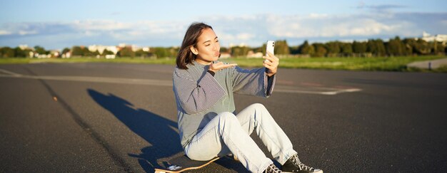 Happy asian girl sits on skateboard takes selfie with longboard makes cute faces sunny day outdoors