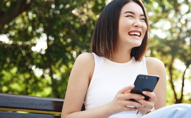 Счастливая азиатская девушка, сидящая на скамейке и использующая мобильный телефон
