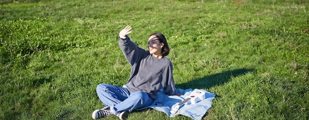 Бесплатное фото Счастливая азиатская девушка сидит на одеяле для пикника с укулеле, прикрывая себя от солнечного света, протягивая руку