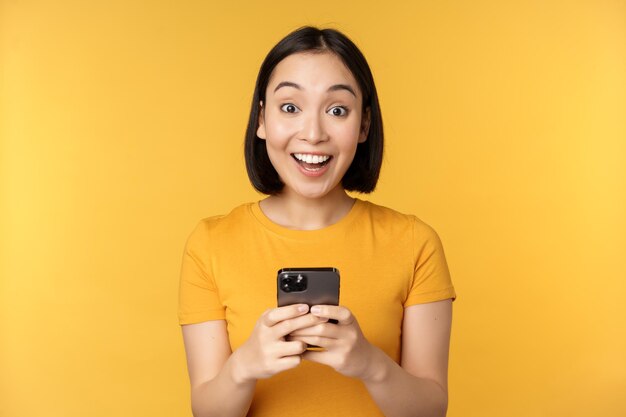 노란색 배경에 검은색 휴대폰을 들고 서서 웃고 있는 행복한 아시아 소녀