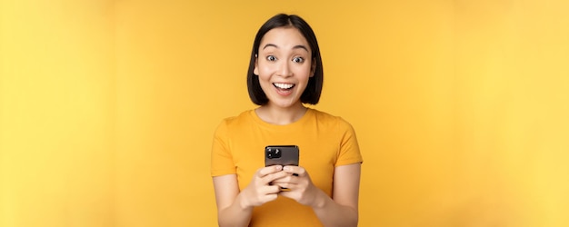 Счастливая азиатская девушка улыбается, стоя с черным мобильным телефоном, стоящим на желтом фоне