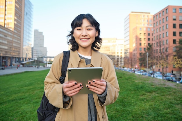 幸せなアジアの女の子が通りに立っている大学生の手でデジタル タブレットと笑顔 s を歩く