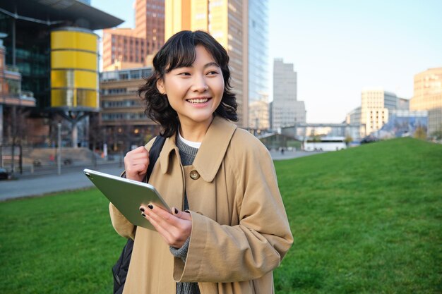 幸せなアジアの女の子が通りに立っている大学生の手でデジタル タブレットと笑顔 s を歩く