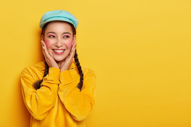 Happy Asian girl touches rouge cheeks, wears makeup and piercing, stylish cap and velvet sweatshirt, has dreamy expression