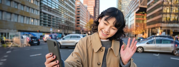 Free photo happy asian girl tourist takes selfie in city centre makes video call and waves at smartphone camera