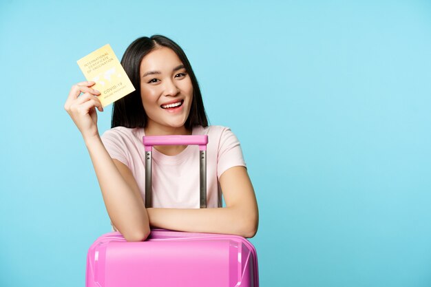 Happy asian girl tourist with suitcase shows her covid international vaccination passport getting va...