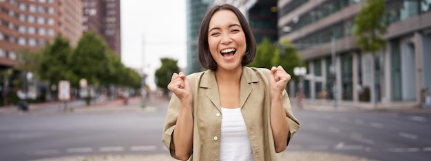 Free photo happy asian girl triumphing on streets of city dancing from happiness celebrating victory posing