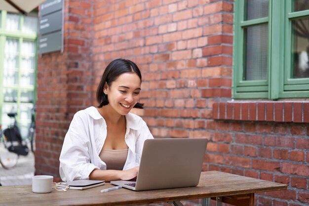 Happy asian girl watching video on laptop doing homework outdoor in coworking space smiling and work