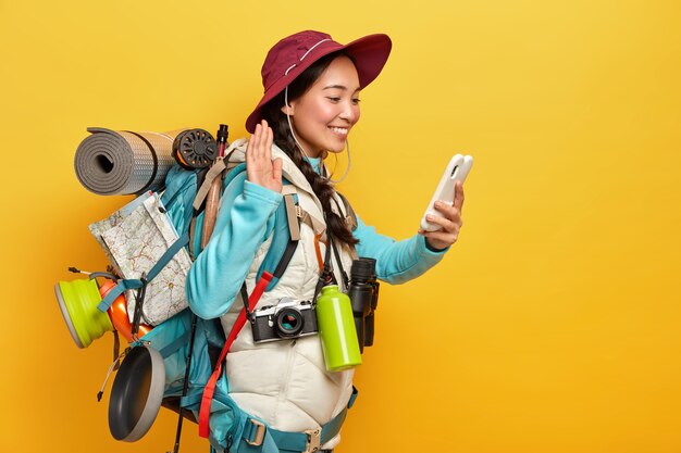 Happy Asian girl waves palm, greets someone via video call, holds mobile phone in hand, carries rucksack with all necessary things, has hiking tour, isolated over yellow wall