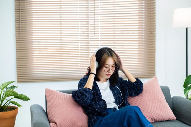 Счастливая азиатская девушка в очках слушает музыку в наушниках в гостиной дома Концепция отдыха дома