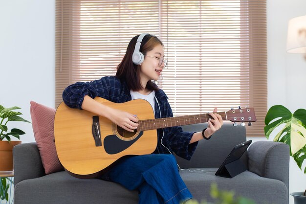 Счастливая азиатская девушка в очках играет на акустической гитаре и слушает музыку с планшетного компьютера в гостиной дома Концепция отдыха дома