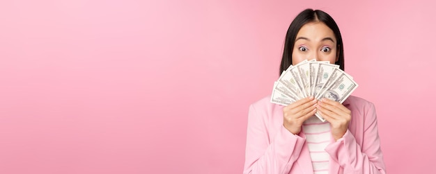 Счастливая азиатка в костюме, держащая деньги в долларах с довольным выражением лица, стоящая над розовым задником