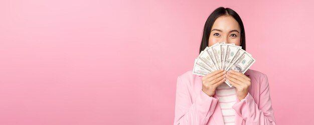 Happy asian lady in suit holding money dollars with pleased face expression standing over pink background Copy space