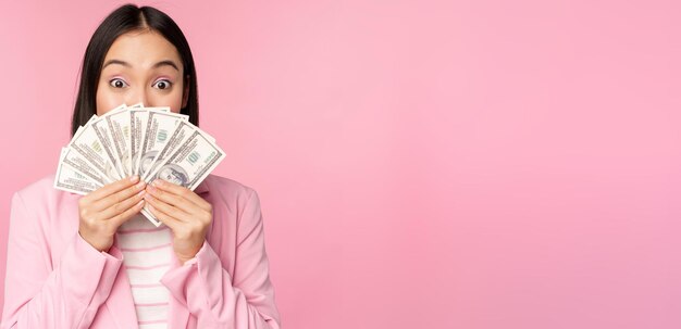 ピンクの背景の上に立っている満足のいく表情でお金のドルを保持しているスーツの幸せなアジアの女性