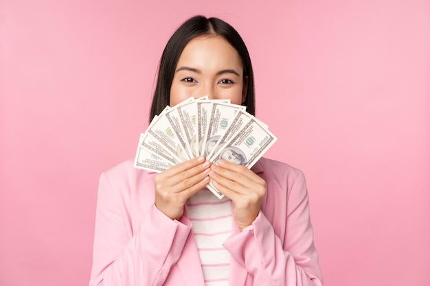 Happy asian lady in suit holding money dollars with pleased face expression standing over pink background