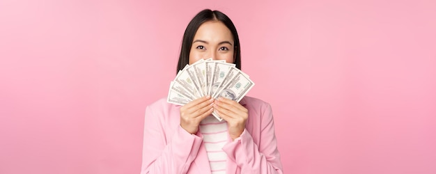 Foto gratuita felice signora asiatica in giacca e cravatta in possesso di dollari di denaro con l'espressione del viso felice in piedi su sfondo rosa