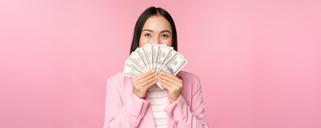 Счастливая азиатка в костюме, держащая деньги в долларах с довольным выражением лица, стоящая на розовом фоне