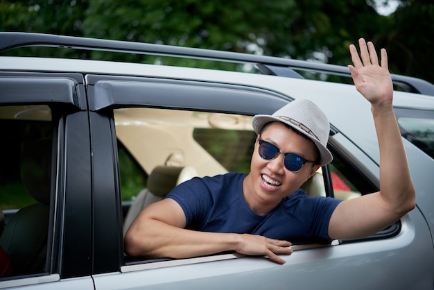 Happy Asian man in hat and sunglasses leaning out of rear window of car and waving