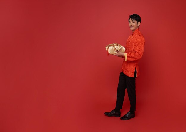 빨간색 전통 의상을 입은 행복한 아시아 남자가 중국 신년 축복을 위해 금 선물 상자를 주고 있습니다.
