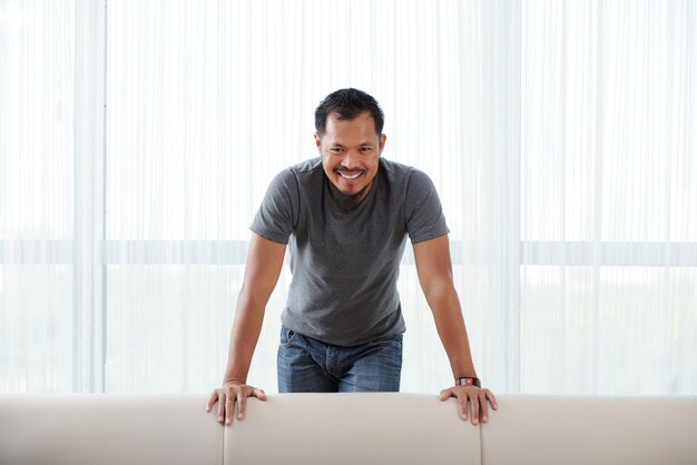 Happy Asian man standing behind couch, leaning on it and smiling for camera 