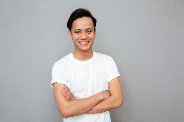 Free photo happy asian man standing with arms crossed over grey wall.