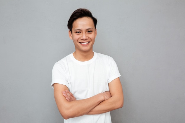 Happy asian man standing with arms crossed over grey wall.