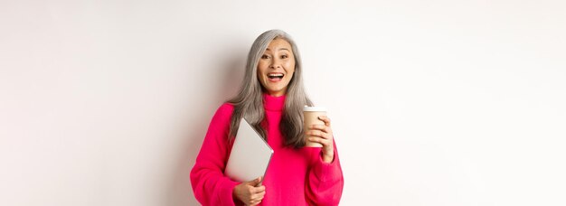 무료 사진 커피를 마시고 분홍색으로 서 있는 노트북을 들고 웃고 있는 행복한 아시아 고위 여성 기업가
