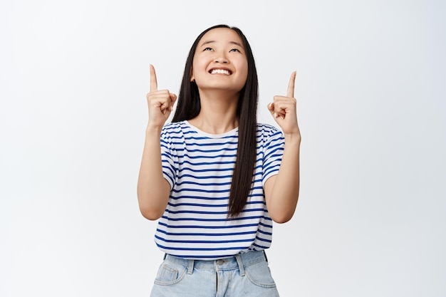 指を上に向けて、白い背景の上にTシャツに立っているトップ広告の夢のような表情を見ている幸せなアジアの10代の少女