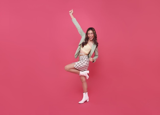 Счастливая азиатка-подросток улыбается и стоит с поднятой рукой, празднуя жест на розовом фоне
