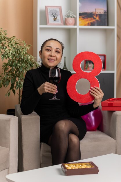 happy asian woman in black dress sitting on a chair with glass of wine holding number eight smiling cheerfully in light living room celebrating international womens day march