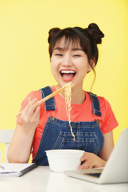 Happy Asian woman carrying instant noodles with chopsticks to mouth, in front of laptop