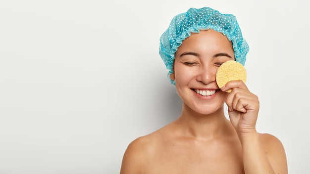 幸せなアジアの女性は化粧用スポンジで顔をきれいにし、化粧を取り除き、目を閉じて、保護シャワーキャップを着用します