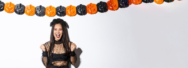 Happy asian woman enjoying halloween wearing wicked witch costume and rejoicing against pumpkin stre