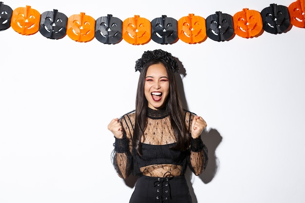 Happy asian woman enjoying halloween, wearing wicked witch costume and rejoicing against pumpkin streamers decoration.