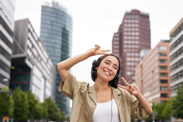 Free photo happy asian woman in headphones listening music and dancing on street of city centre smiling with ha