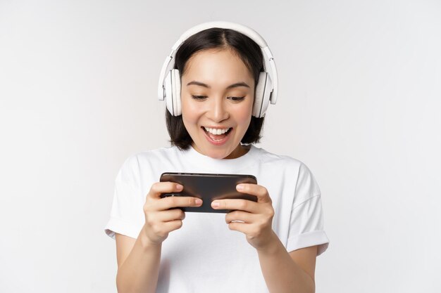 Happy asian woman in headphones looking at smartphone watching video on mobile phone and smiling standing over white background