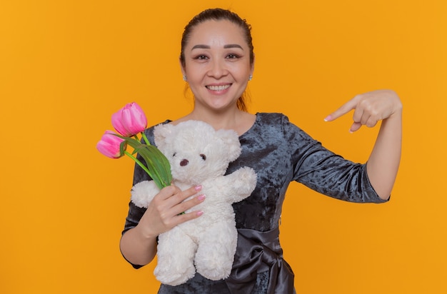 Free photo happy asian woman holding bouquet of pink tulips and teddy bear pointing