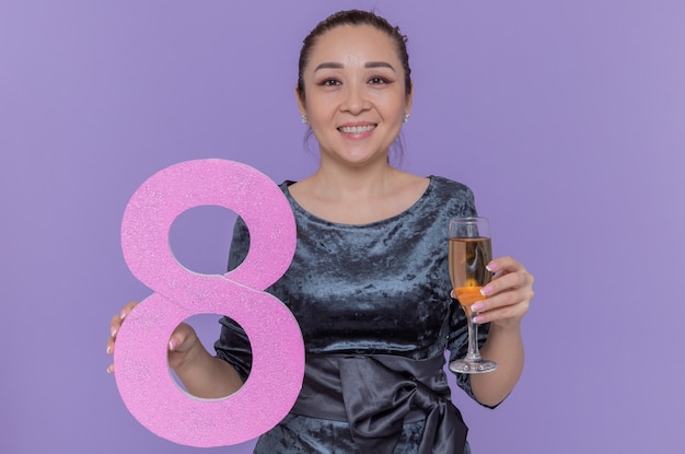 Foto gratuita felice donna asiatica tenendo il numero otto realizzato in cartone e un bicchiere di champagne guardando davanti sorridendo allegramente per celebrare la giornata internazionale della donna in piedi sopra la parete viola