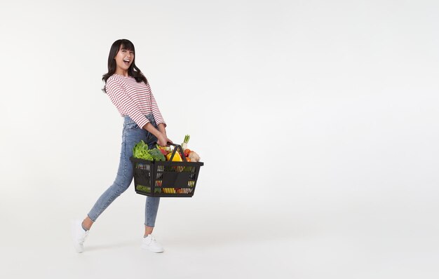 Счастливая азиатка с корзиной для покупок, полной овощей и продуктов, снятая в студии